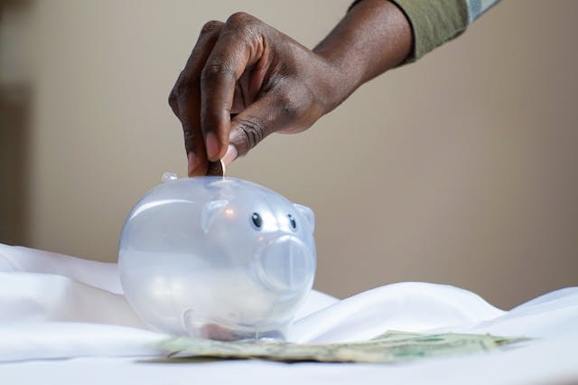 hand-placing-coin-into-transparent-piggy-bank-with-money-below